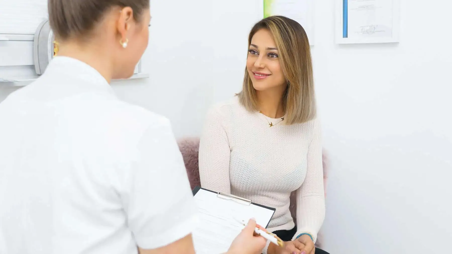 Woman in white sweater smiling and talking to a healthcare professional holding a clipboard.