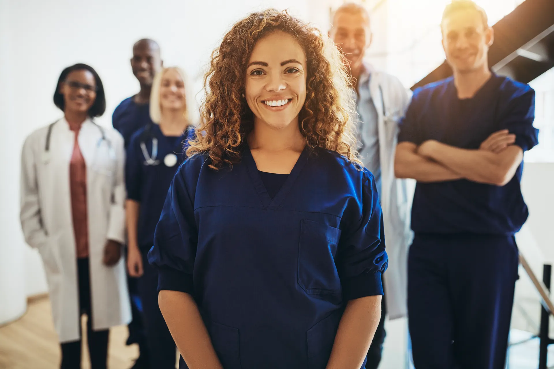 Smiling female nurse in blue scrubs stands front of a diverse medical team in a bright hospital.
