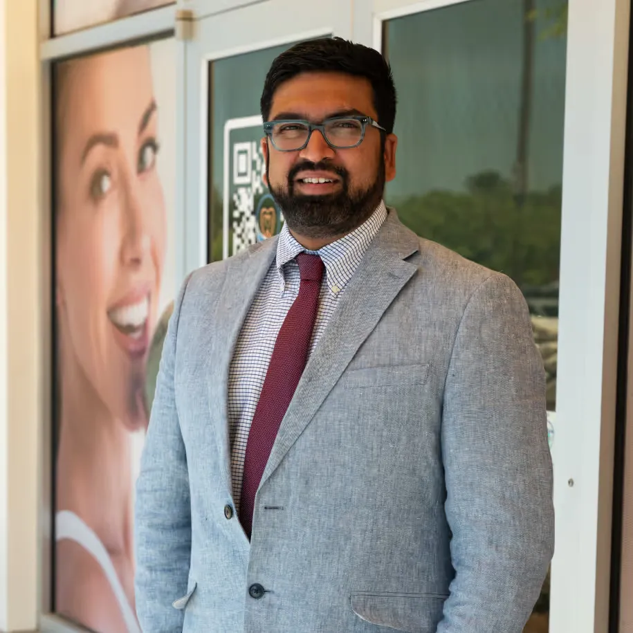 Man in glasses, gray blazer, and red tie smiling outside a glass door with an advertisement poster behind.