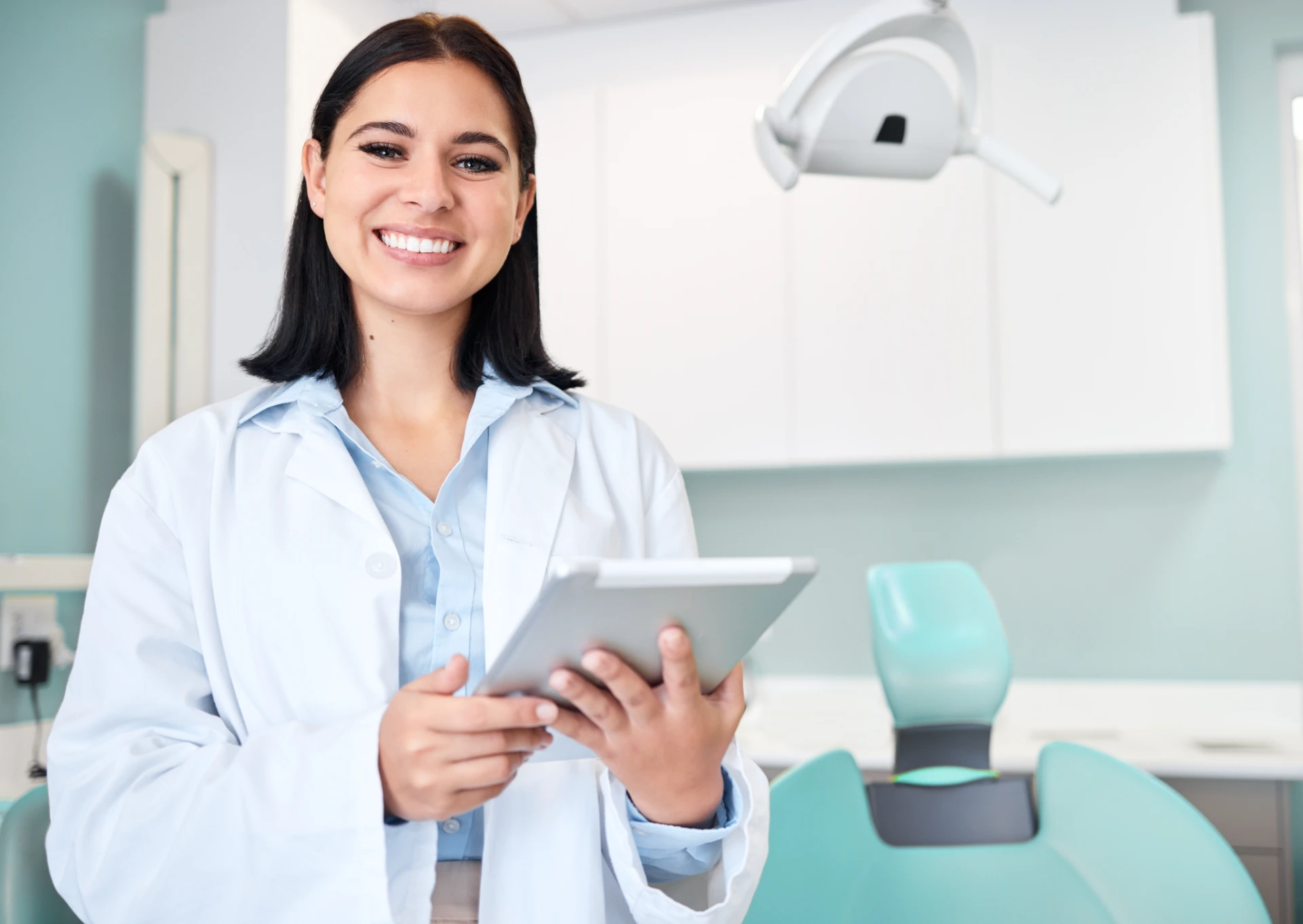 Smiling female dentist in white coat holding a tablet in a dental office with a chair and light behind her.