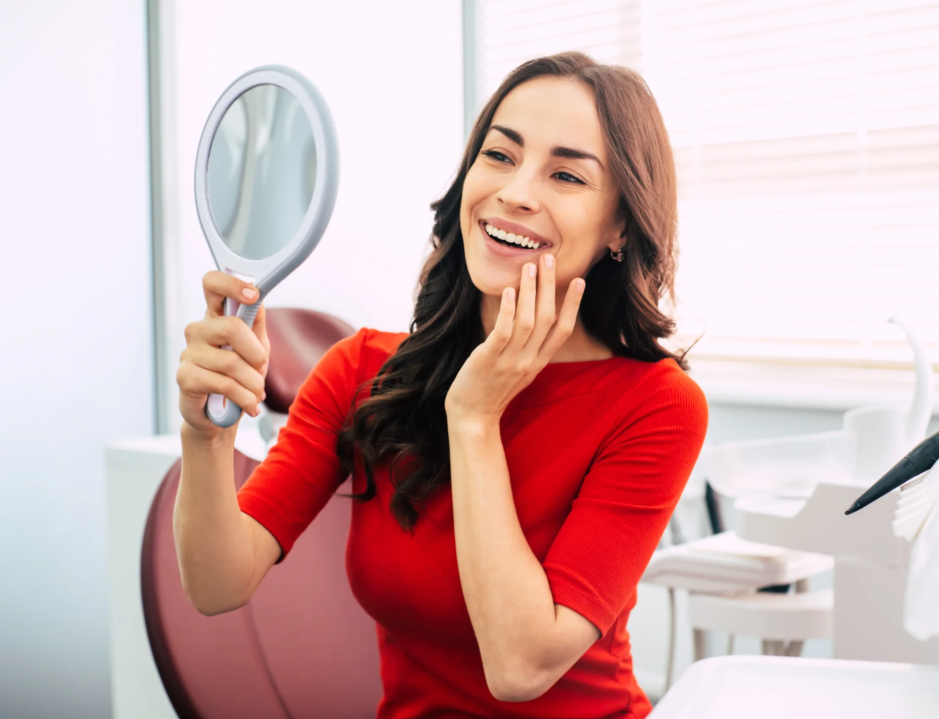 Smiling woman in red shirt holding a handheld mirror, checking her teeth in a dental clinic.