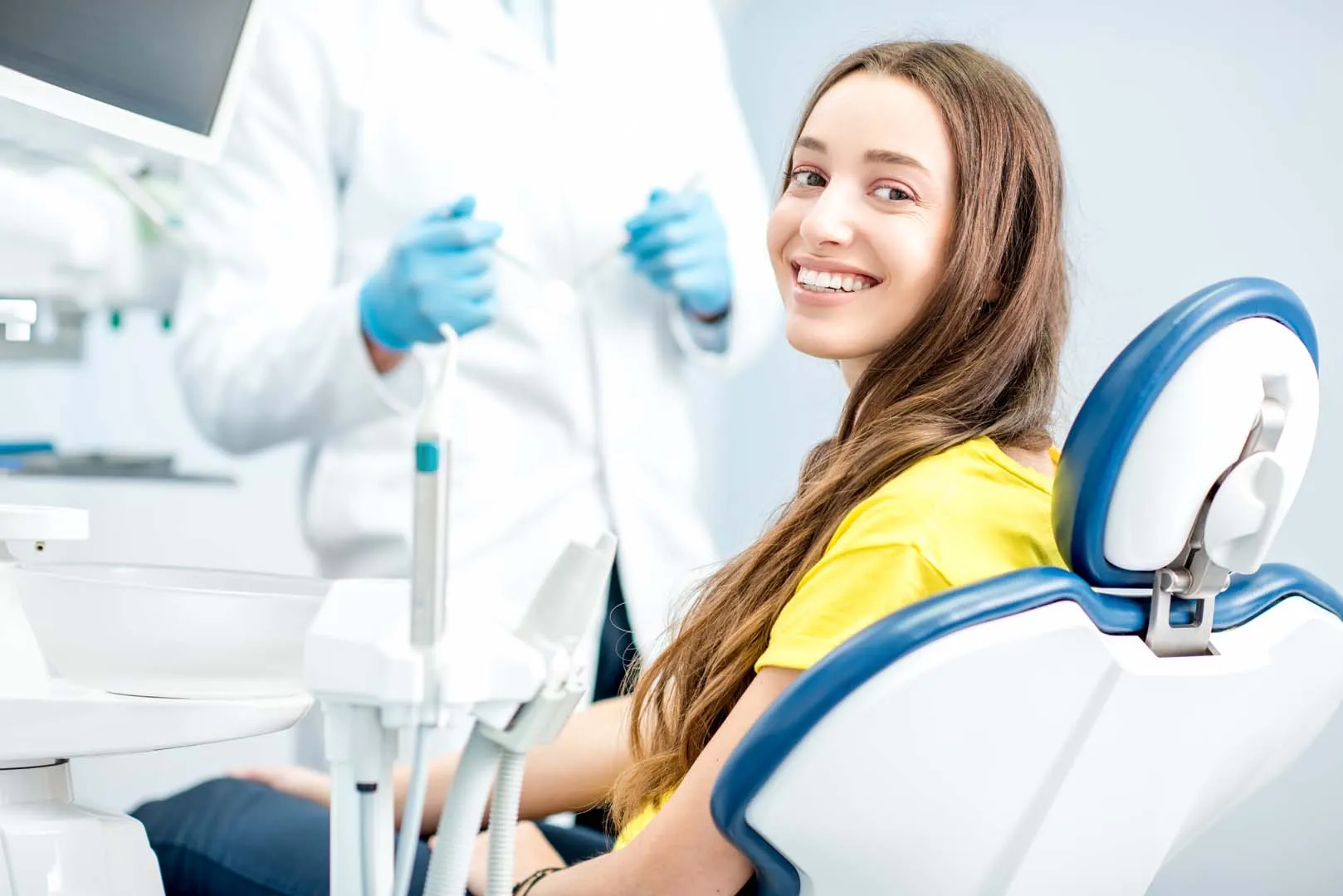 Smiling young woman sitting in a dental chair with a dentist in the background wearing blue gloves.