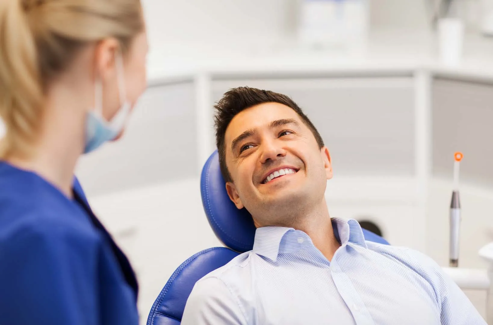 Smiling man in a dental chair looking at a female dentist wearing a mask and blue scrubs.