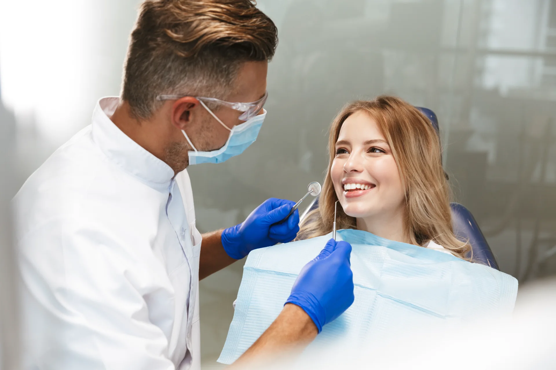 Dentist in protective gear examining smiling woman's teeth in dental clinic.