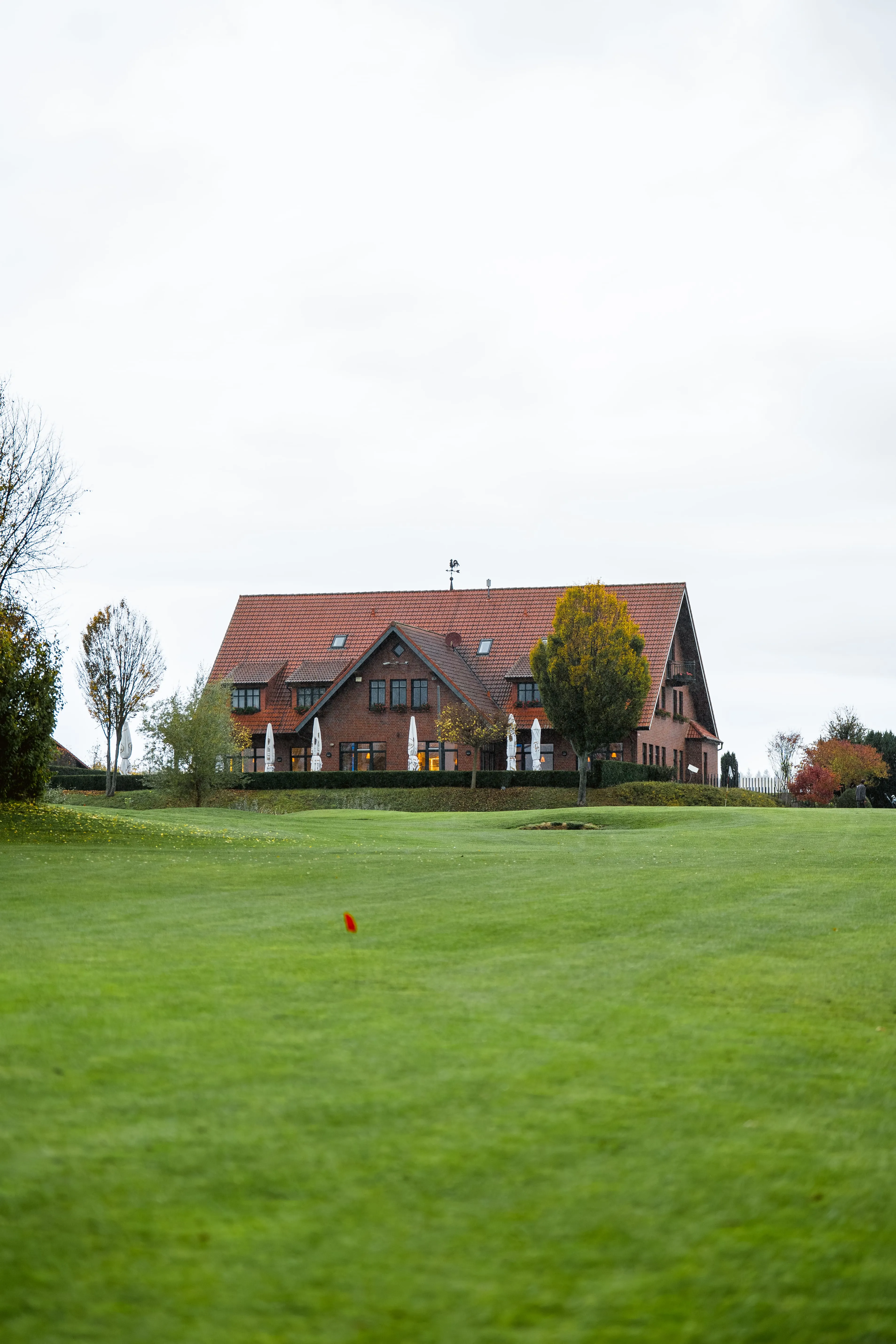 Blick über das Fairway auf das Golfhaus am Löwenhof mit geschlossenen Sonnenschirmen auf der Terrasse
