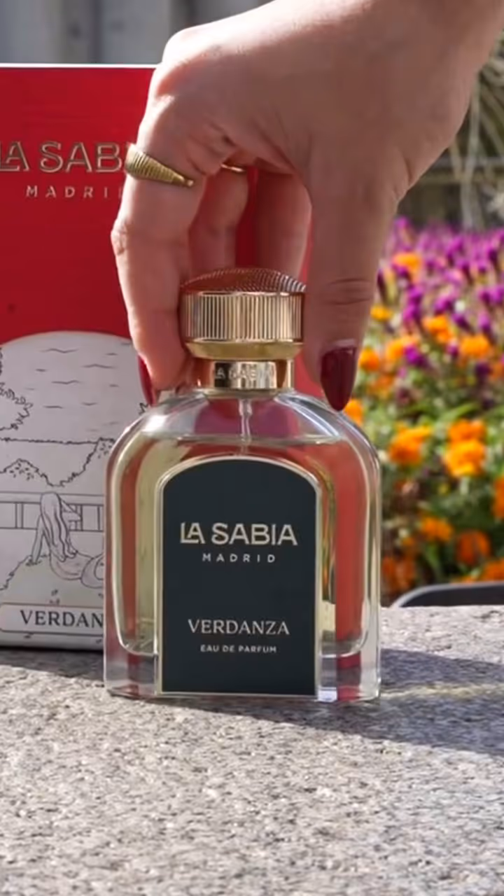 Hand holding La Sabia Madrid Verdanza Eau de Parfum bottle with flowers in background.