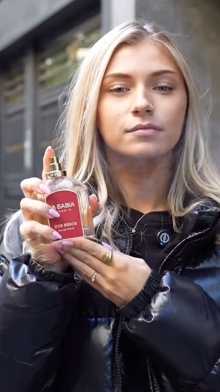 Blonde woman in black jacket holding a perfume bottle labeled La Sabia Dos Besos.