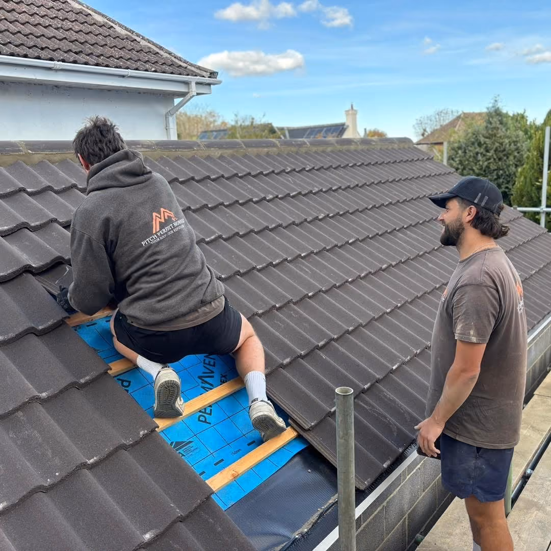 One man kneeling on a roof working on tiles as another watches, under a clear blue sky.