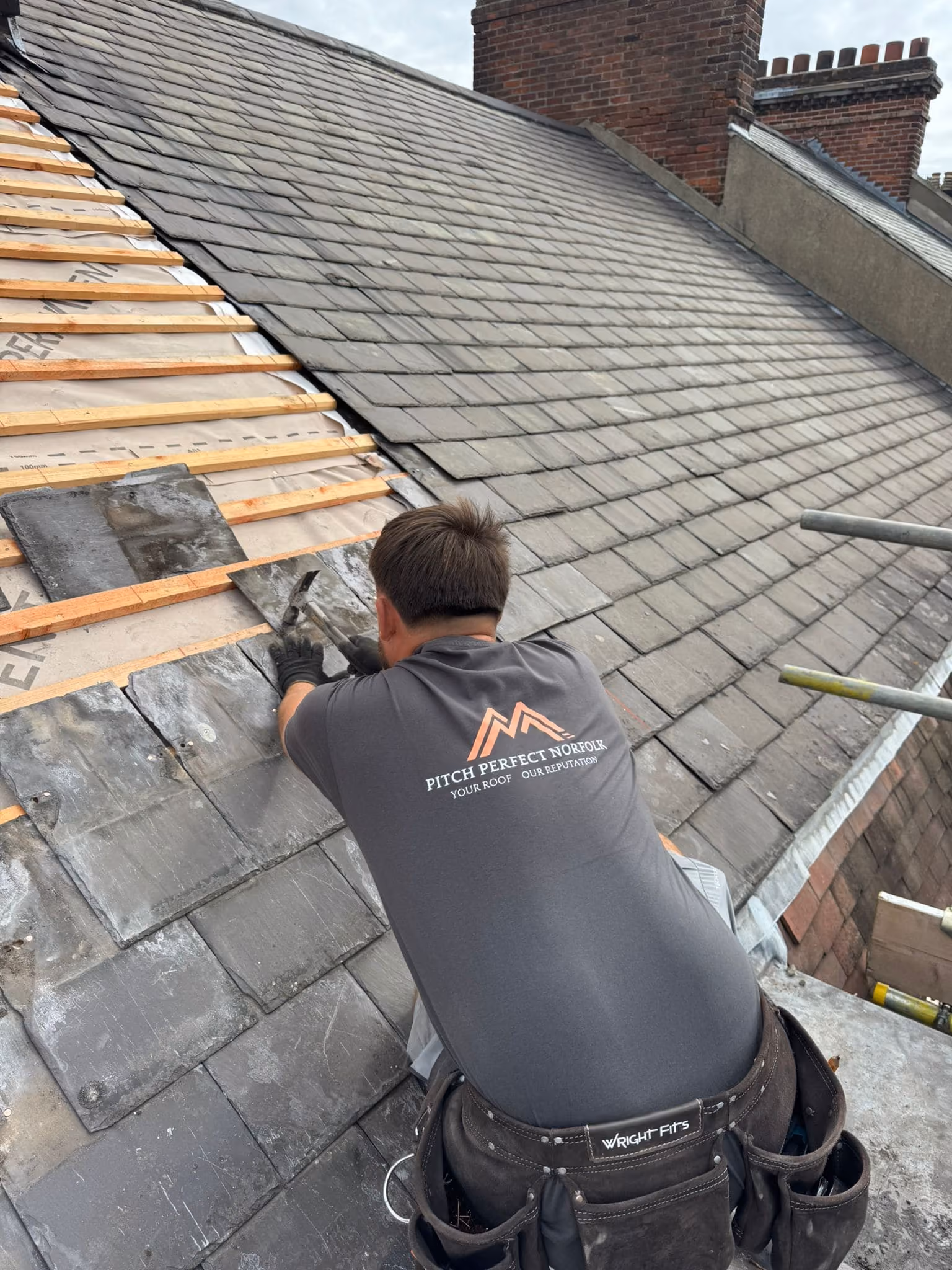 Roofer with Pitch Perfect Norfolk shirt repairing a slate roof using hammer and slate tiles.