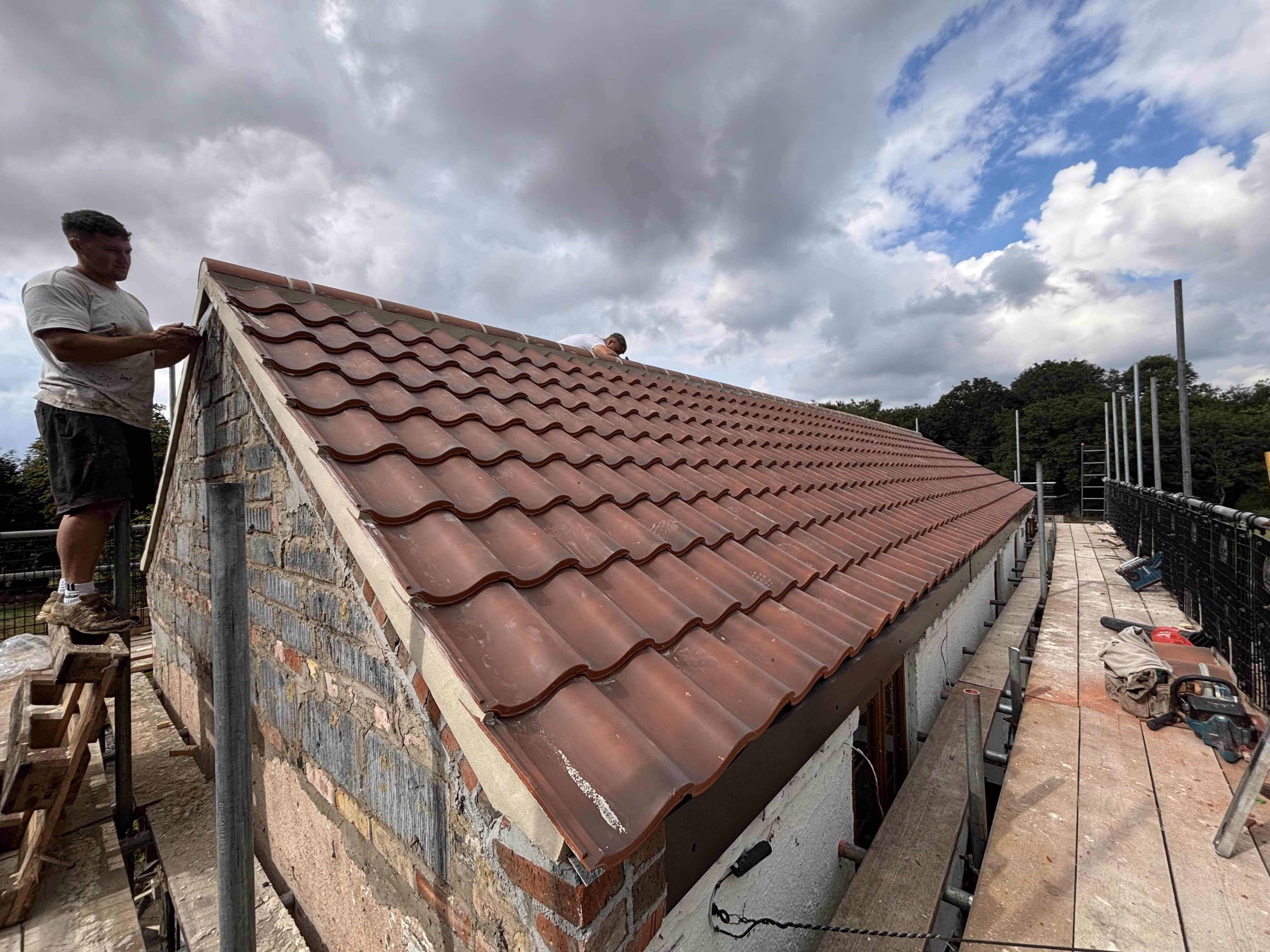 Two construction workers installing brown roof tiles on a building under a cloudy sky.