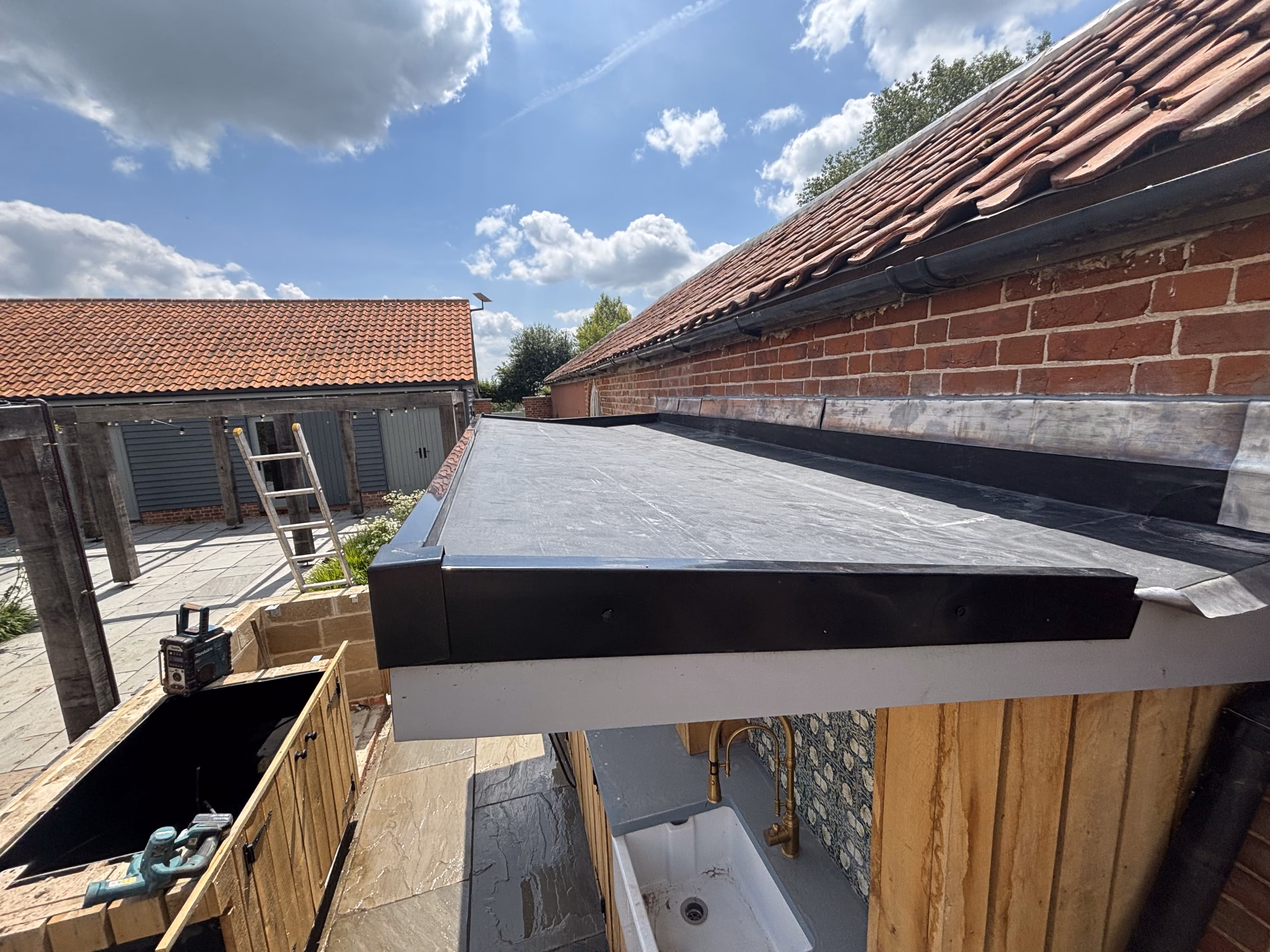 Outdoor area with a flat roof over a sink, wooden cabinetry, a ladder, and partly cloudy sky.