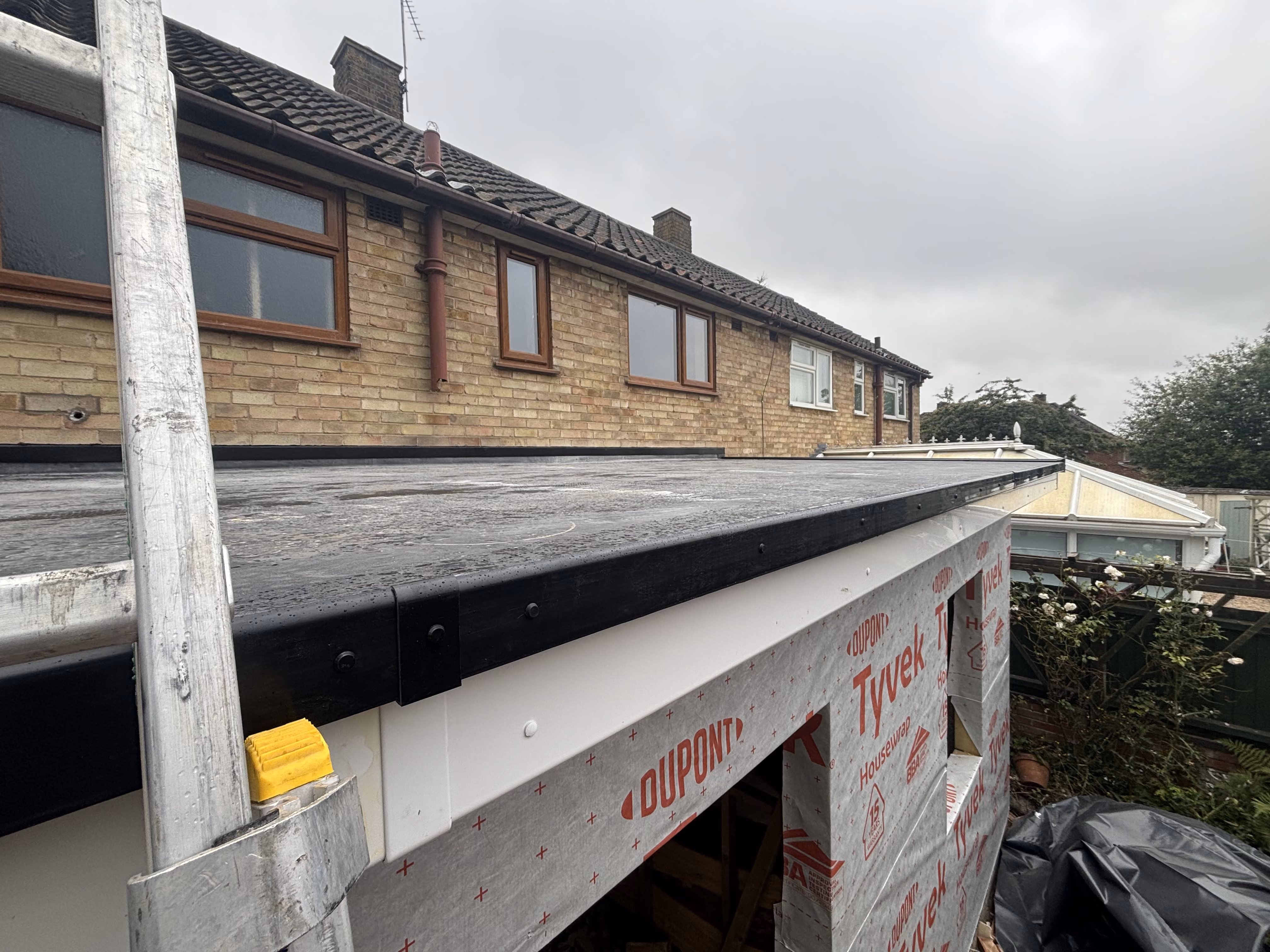 New flat roof with black edging on a house extension under cloudy sky with ladder on the left.