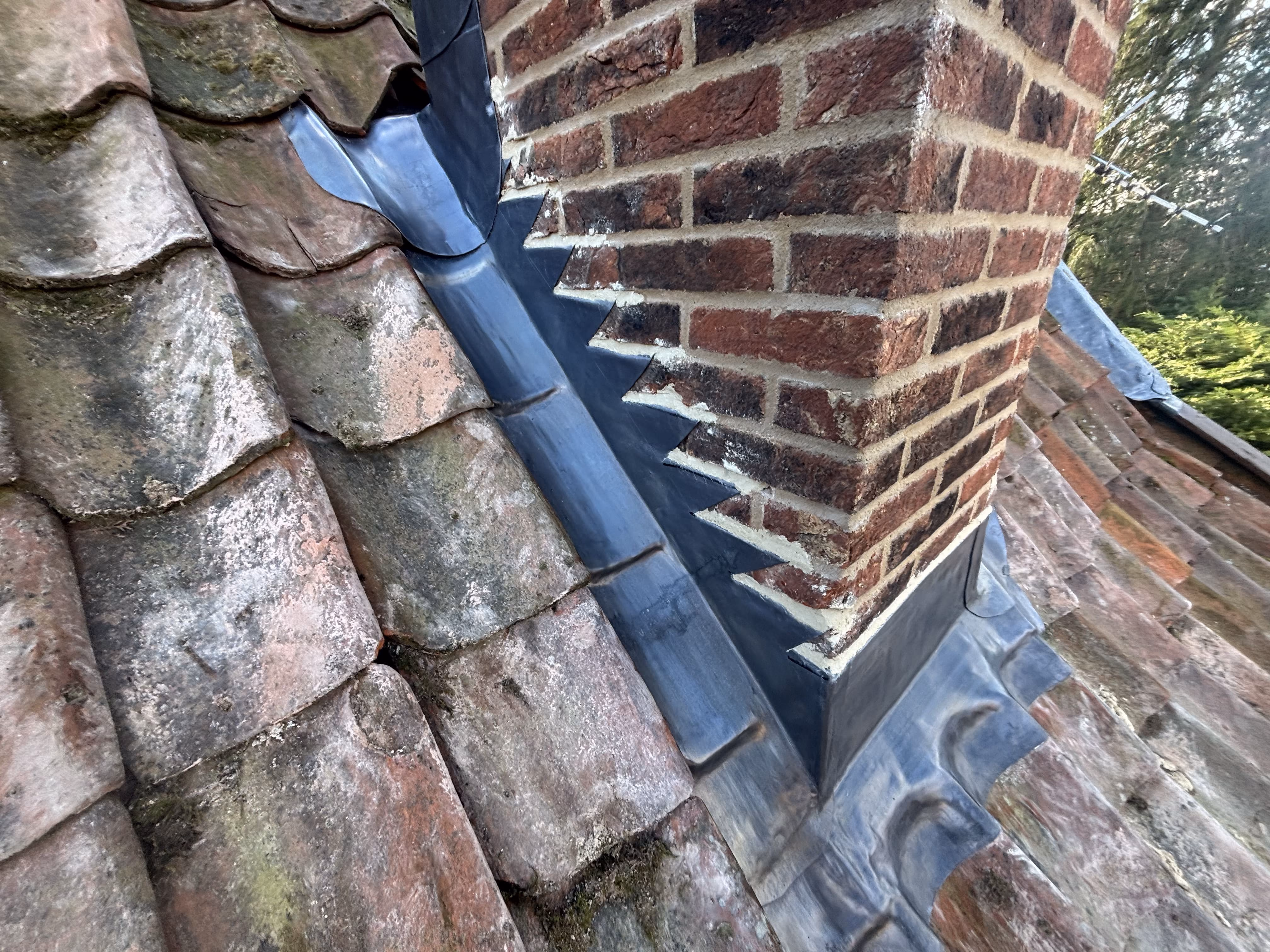 Brick chimney with ornate metal flashing on weathered clay tile roof outdoors.