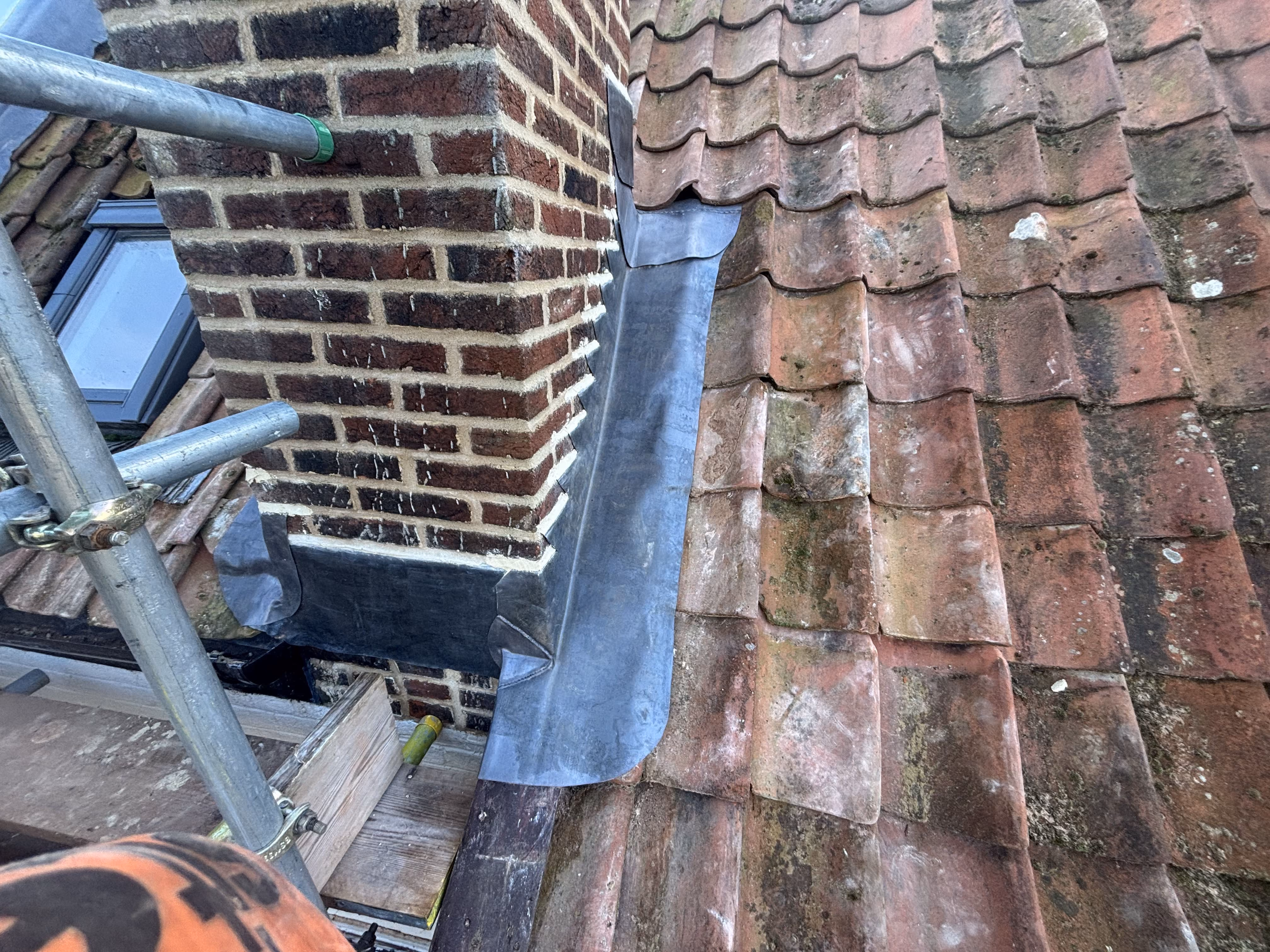 Brick chimney on a tiled roof with metal flashing installed around its base for waterproofing.
