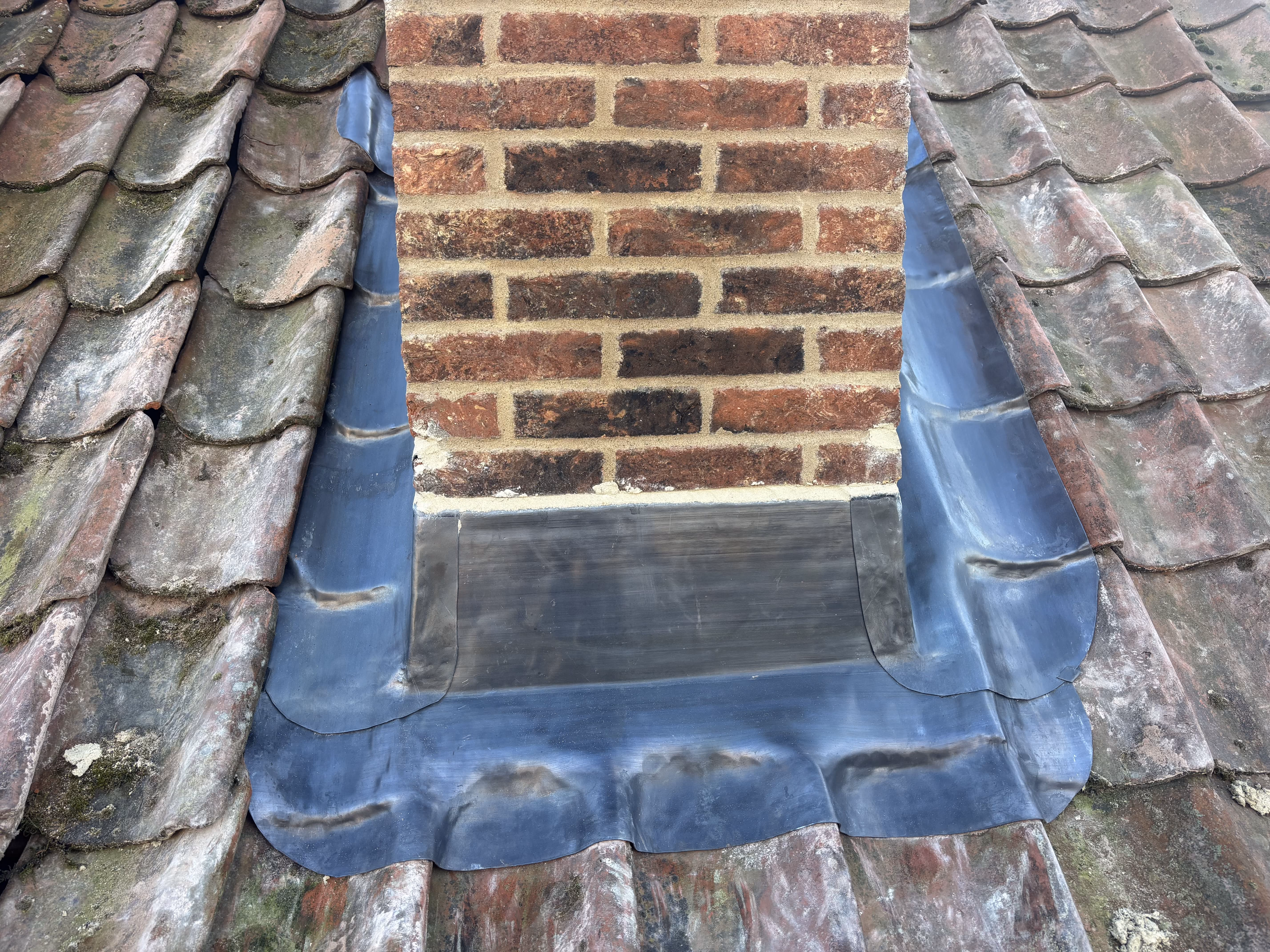 Brick chimney on a roof with old tiles and new black flashing around the chimney base.