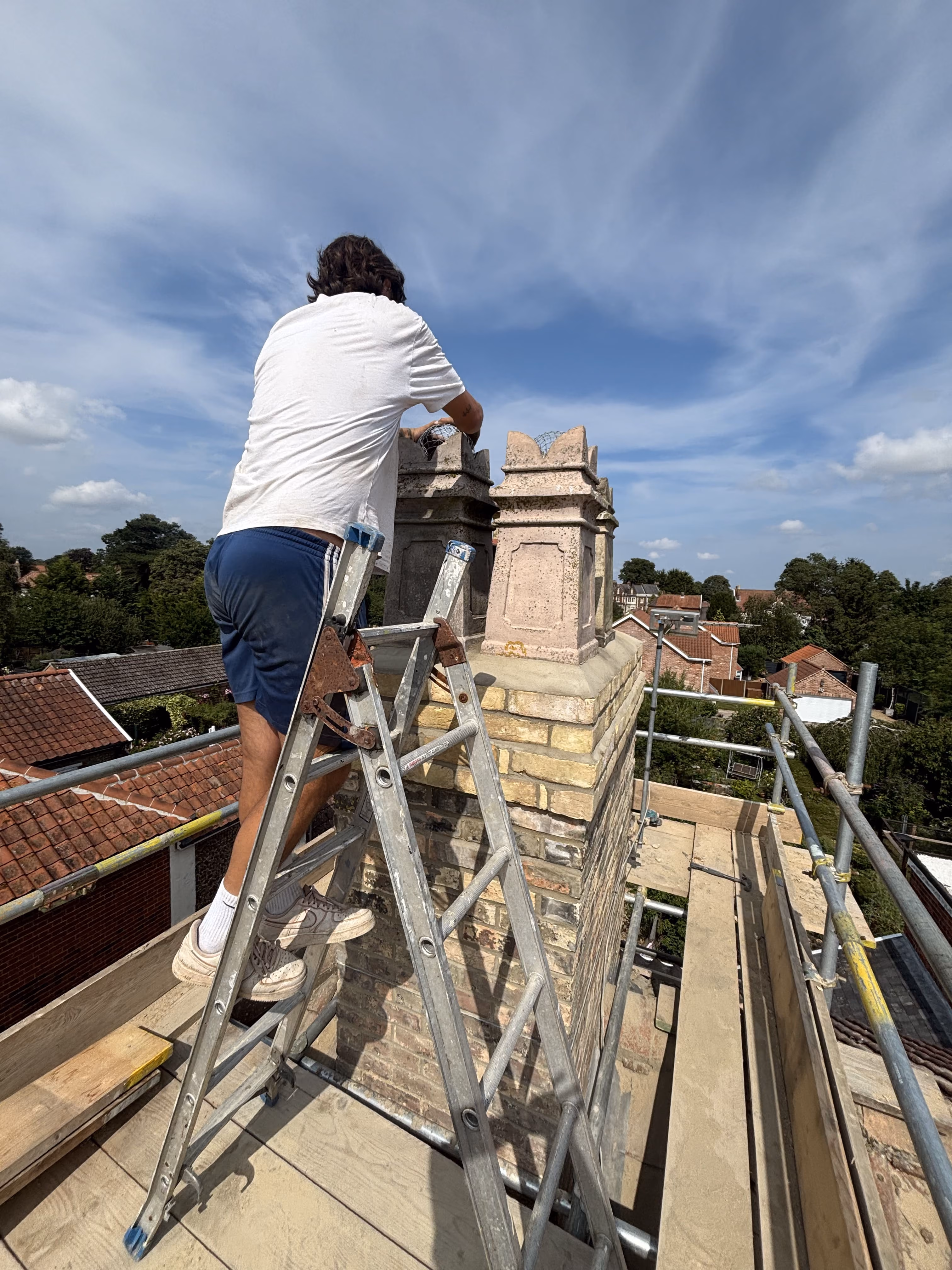 Person on ladder working on brick chimney tops on a roof under blue sky with scattered clouds.