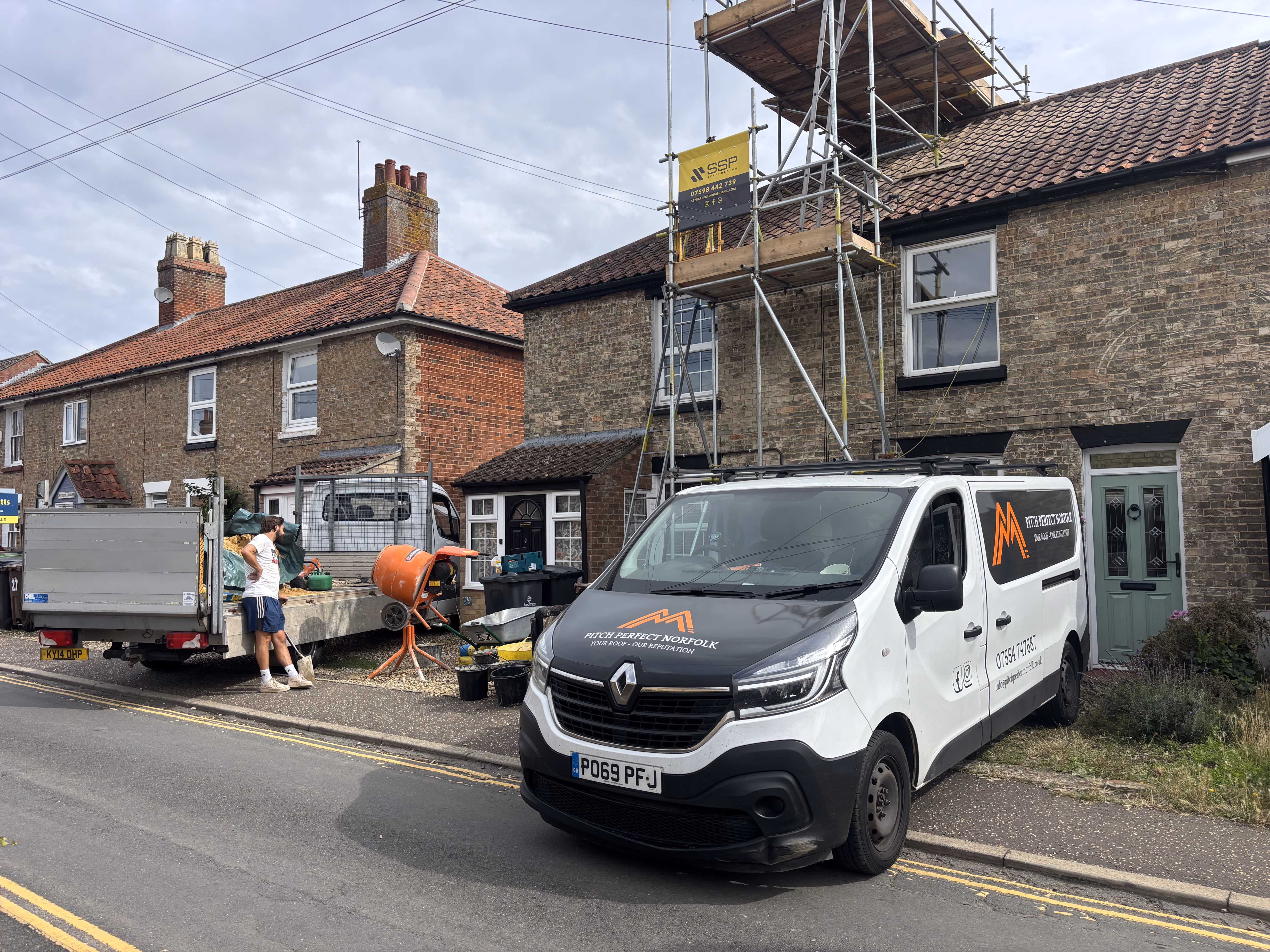 Construction van and worker outside brick house with scaffolding and cement mixer on street.