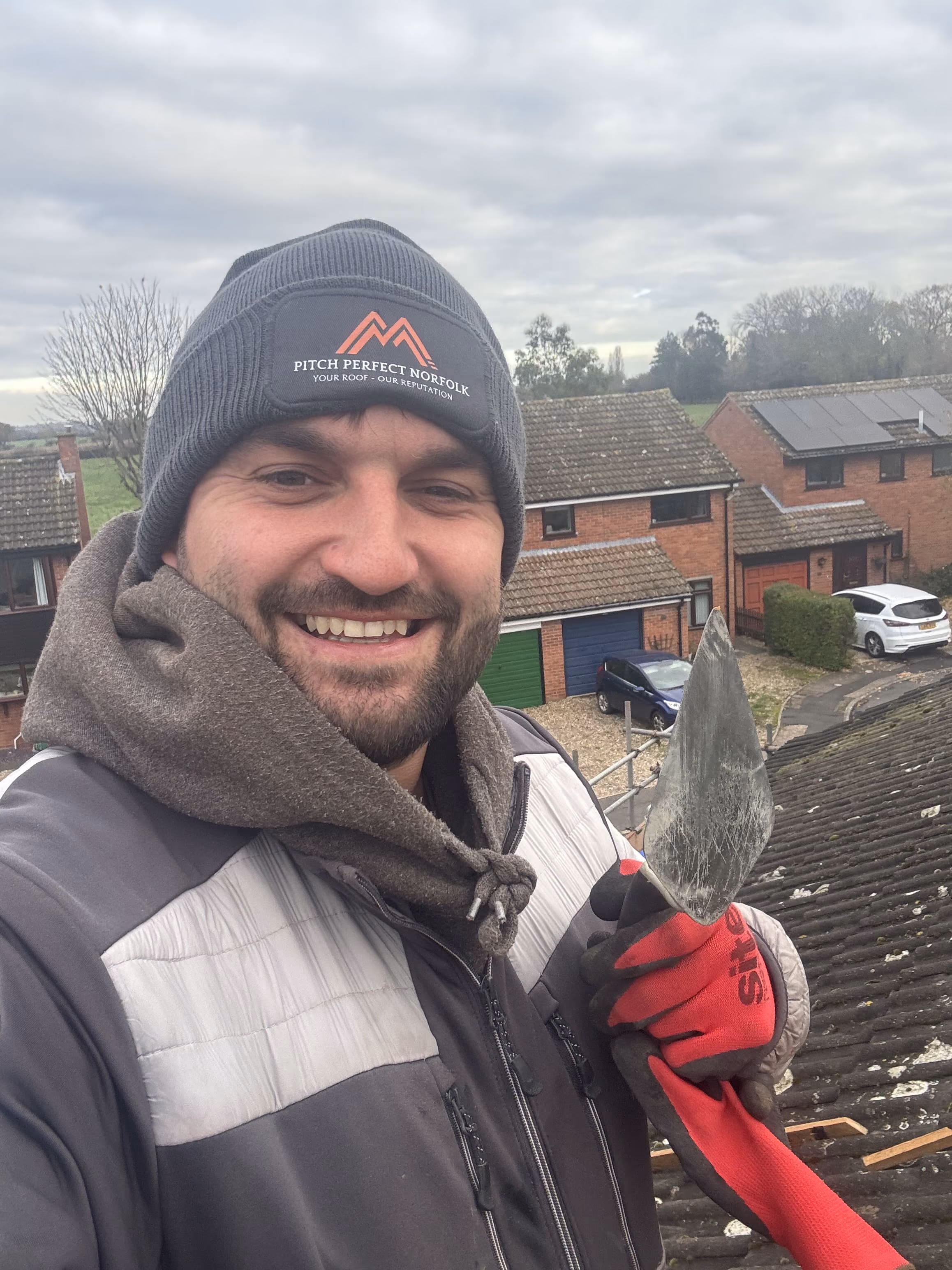 Smiling man wearing a beanie holds a trowel on a rooftop with houses in the background.