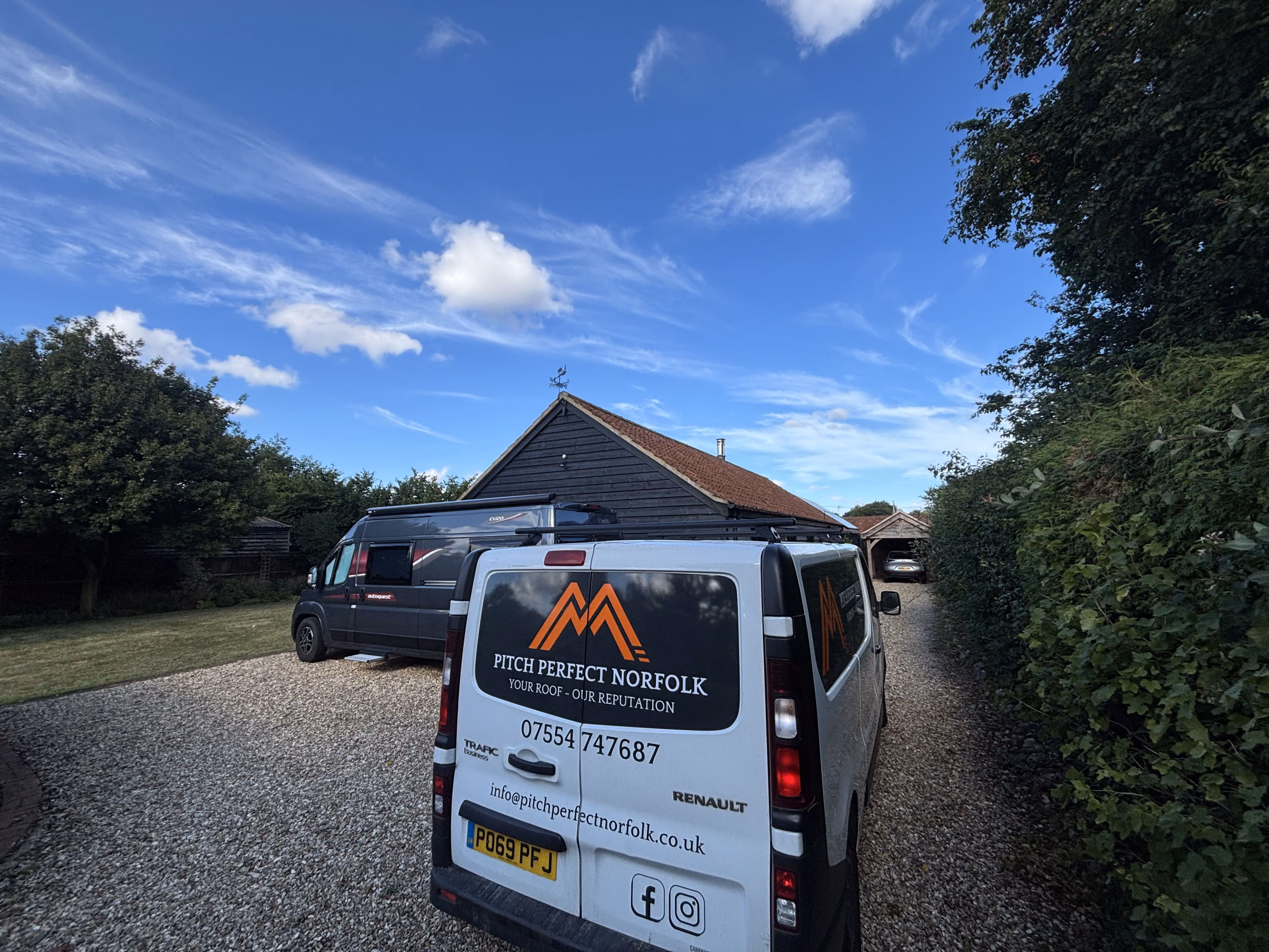 Two vans parked on gravel driveway beside a dark wooden house under a partly cloudy sky.
