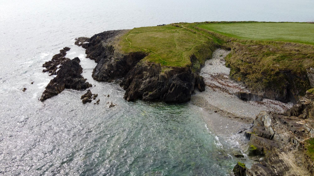 Rocky coastline with grassy cliffs and a small pebble beach by calm ocean waters.