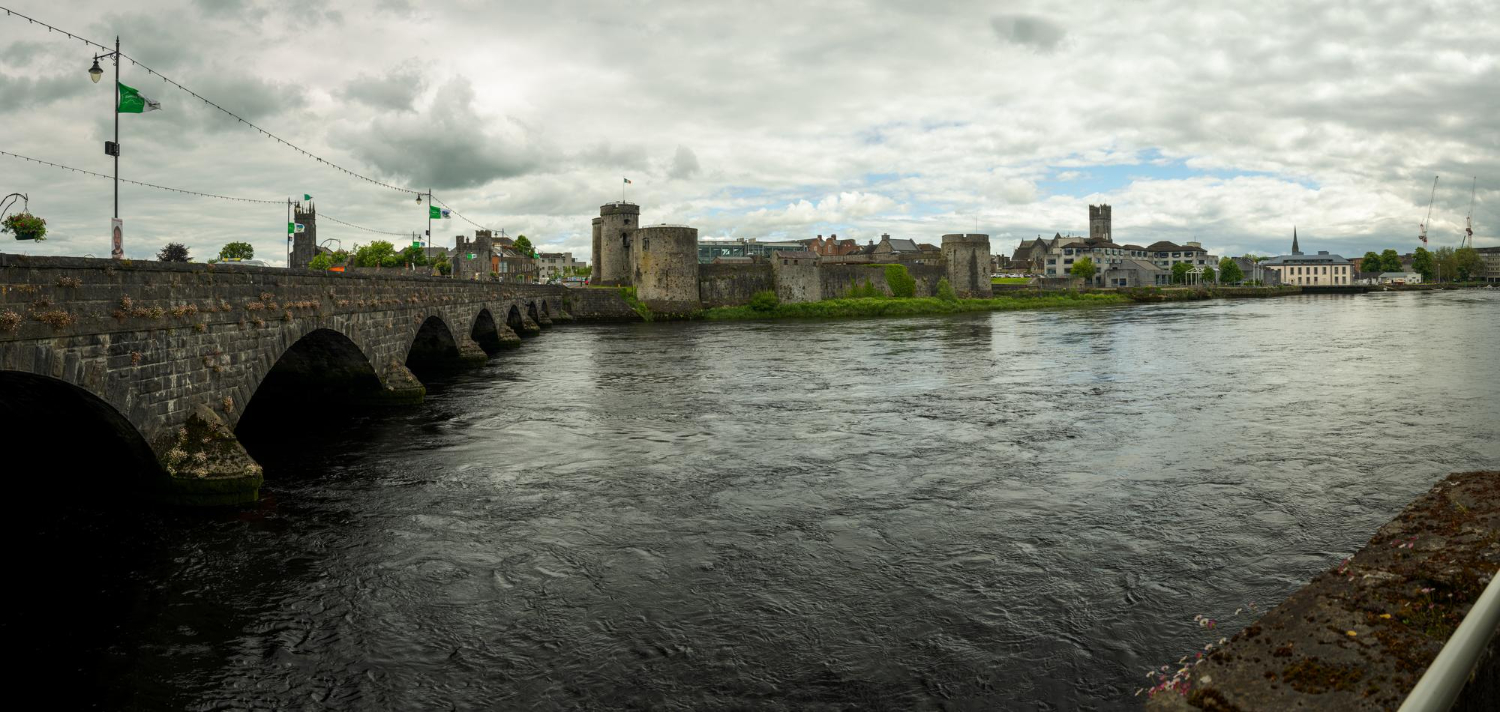 Stone bridge over river with medieval castle and cloudy sky in the background.