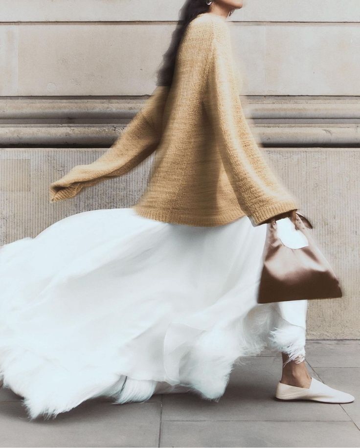 Person walking wearing a tan sweater, long flowing white skirt, beige flats, and carrying a brown handbag.