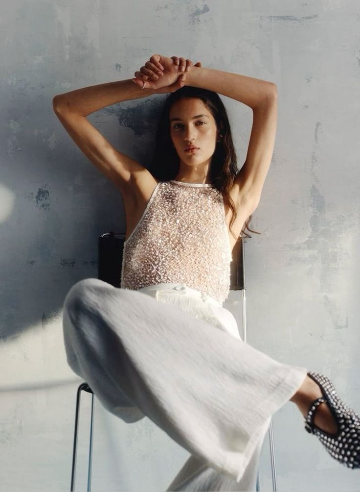 Woman in white textured top and wide pants seated with arms raised against a gray wall.