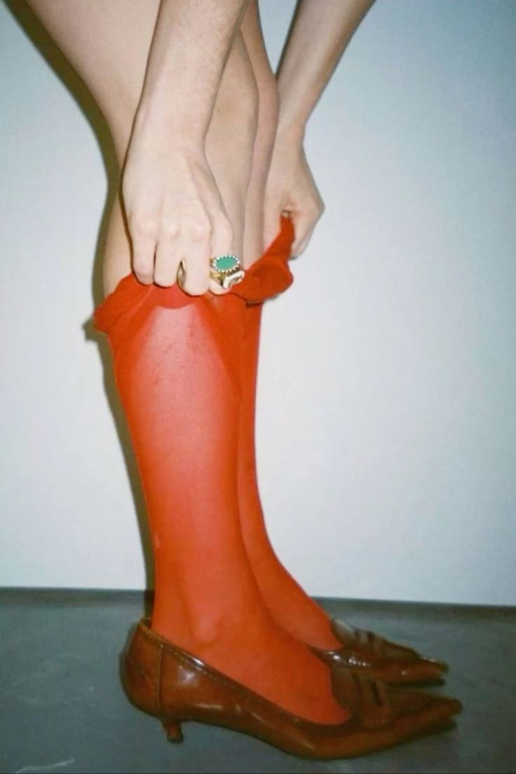 Person pulling up sheer red stockings while wearing brown low-heeled shoes and a large ring.