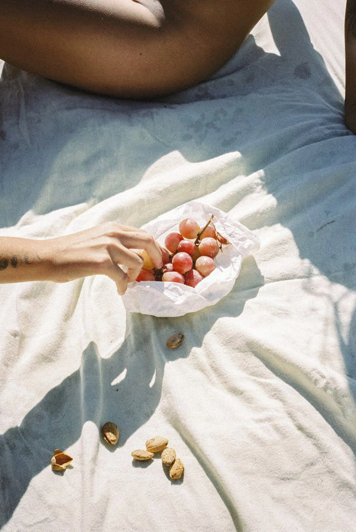 Hand reaching for grapes in a white cloth bowl on a sunlit fabric with scattered nuts.