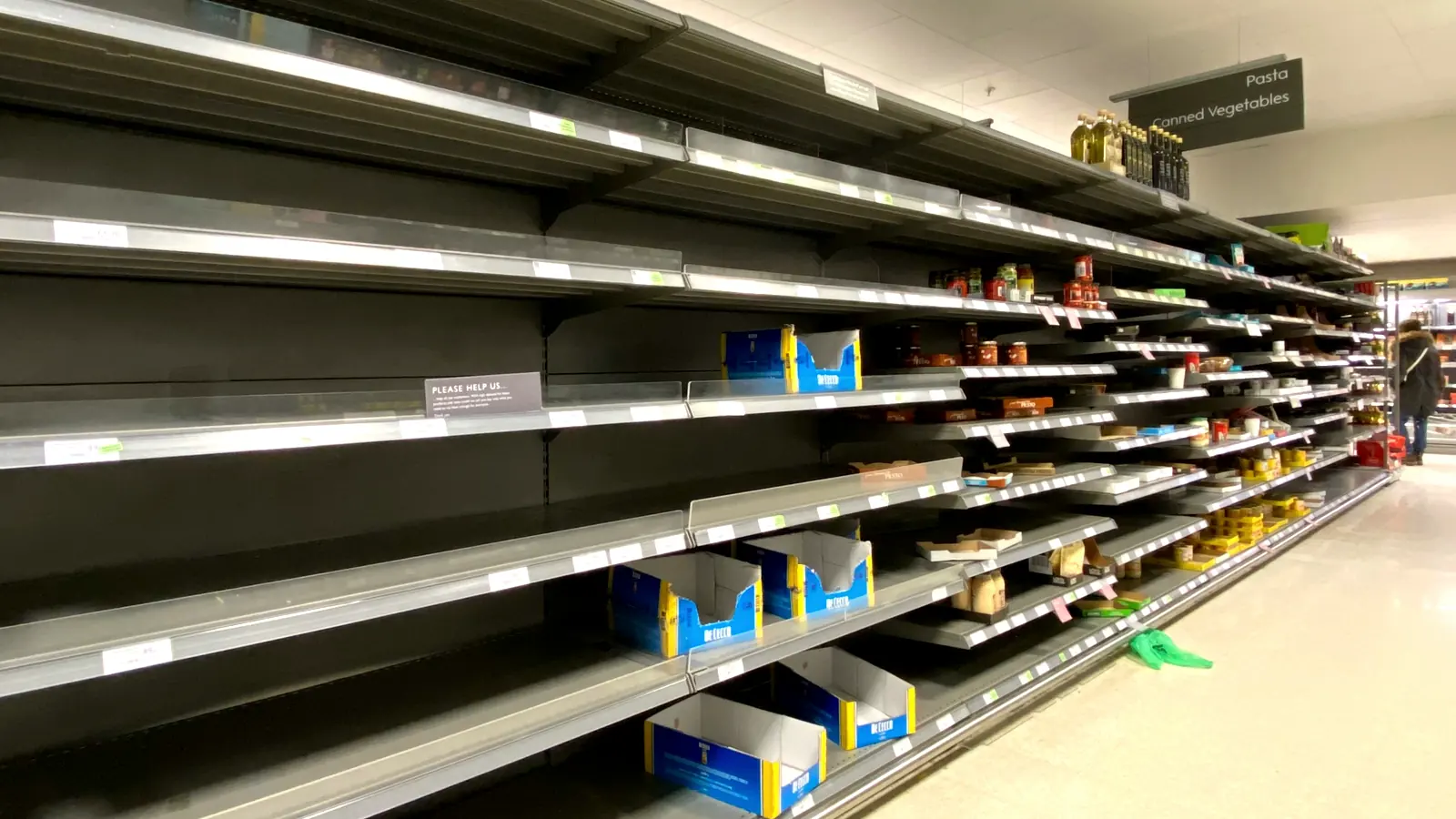 Empty grocery store shelves down both sides of an aisle while a worker restocks from a hand truck of boxes.