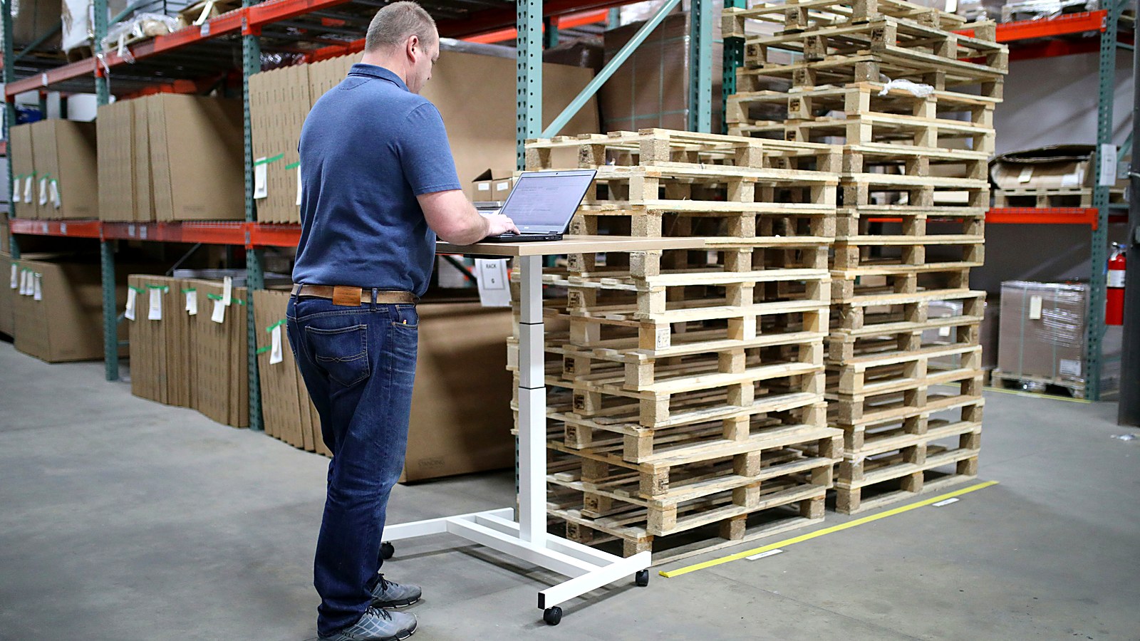Logistics manager reviewing freight rates on a laptop at a standing desk beside stacked pallets in a warehouse