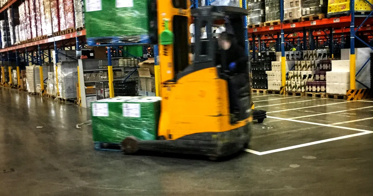 Reach truck moving a stretch-wrapped pallet of cases through racking aisles in a distribution warehouse.