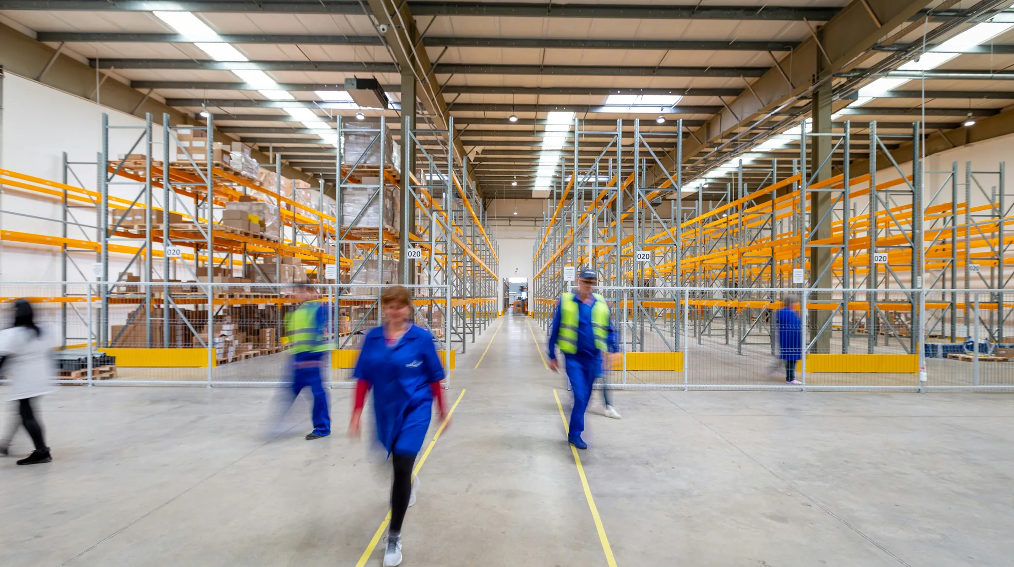 Warehouse workers walking through a distribution center aisle lined with tall pallet racking