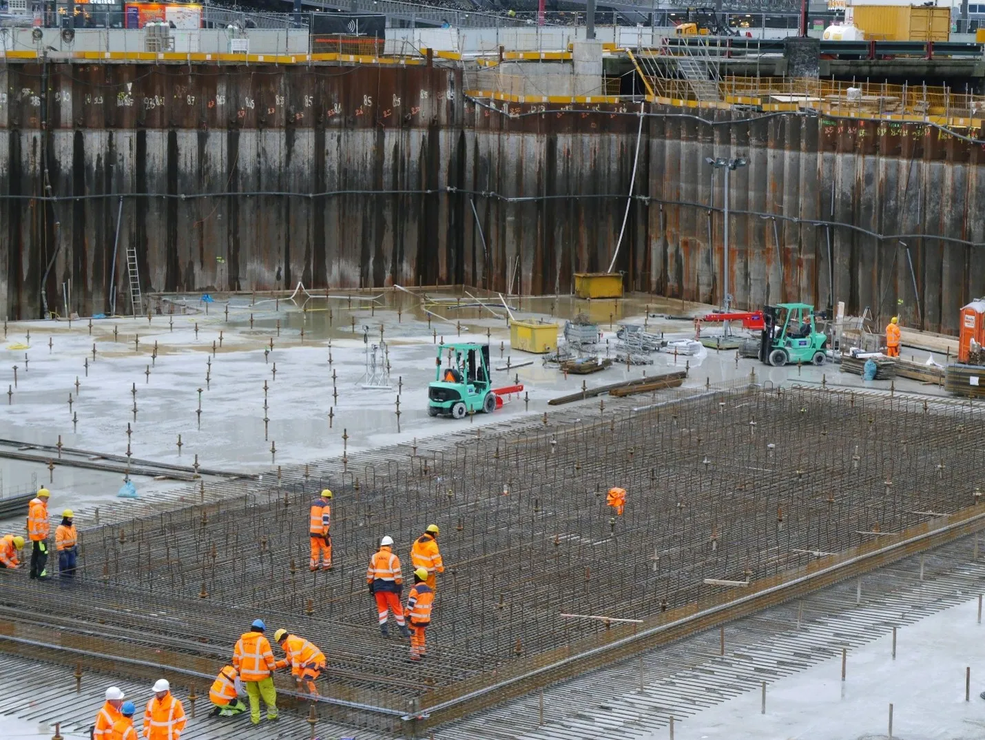 Workers placing steel reinforcement mesh across a large commercial slab inside a shored excavation