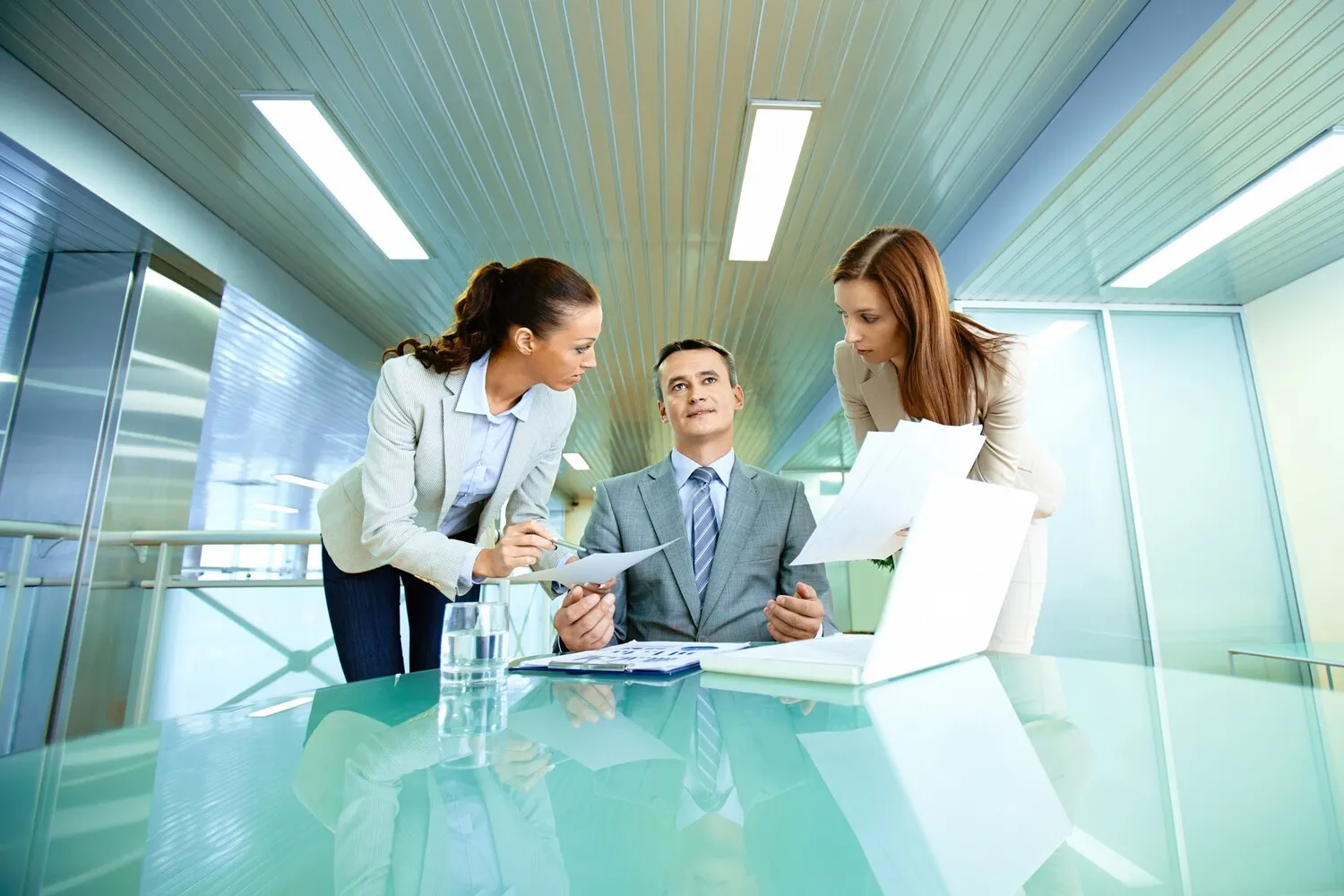 Two businesswomen showing documents to a seated businessman in a modern office meeting room.