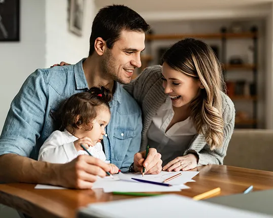 Smiling parents and toddler drawing together with colored pencils at a wooden table at home.