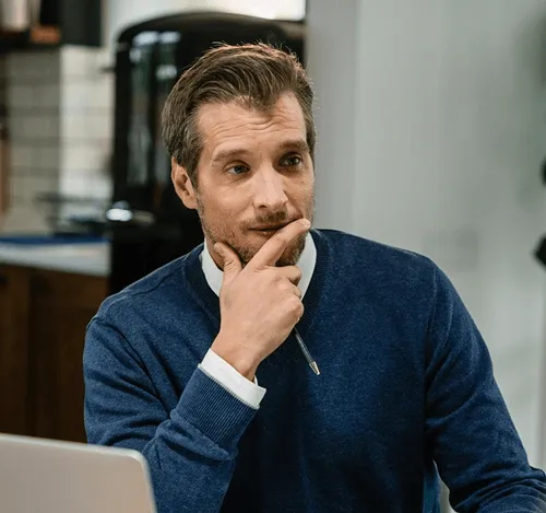 Man in blue sweater holding a pen to his chin, looking thoughtful while seated indoors.
