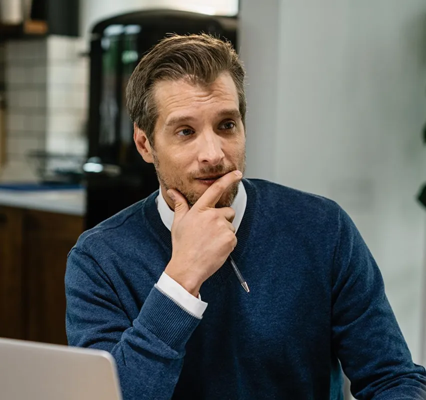 Man in blue sweater holding a pen to his chin, looking thoughtful while seated indoors.