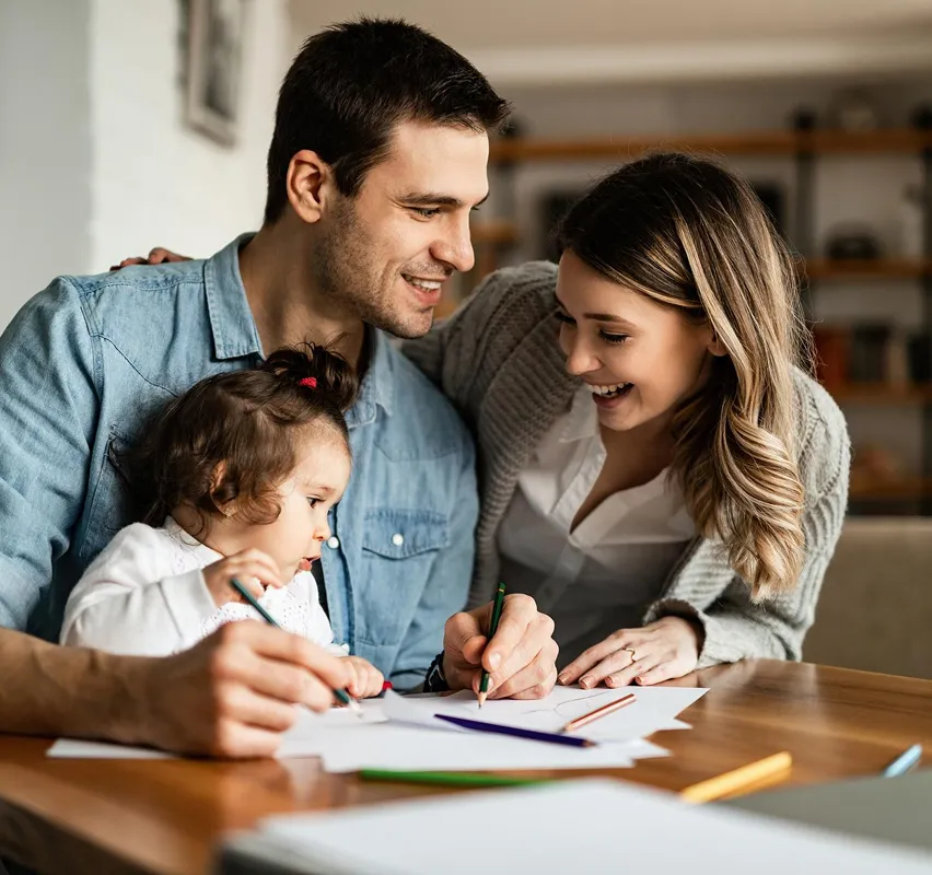 Smiling parents drawing with their toddler daughter at a table covered with paper and colored pencils.