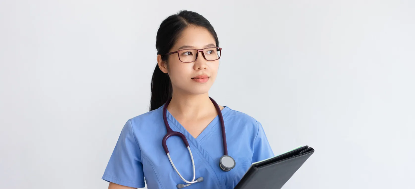Female medical professional in blue scrubs with stethoscope and folder looking thoughtfully to the side.