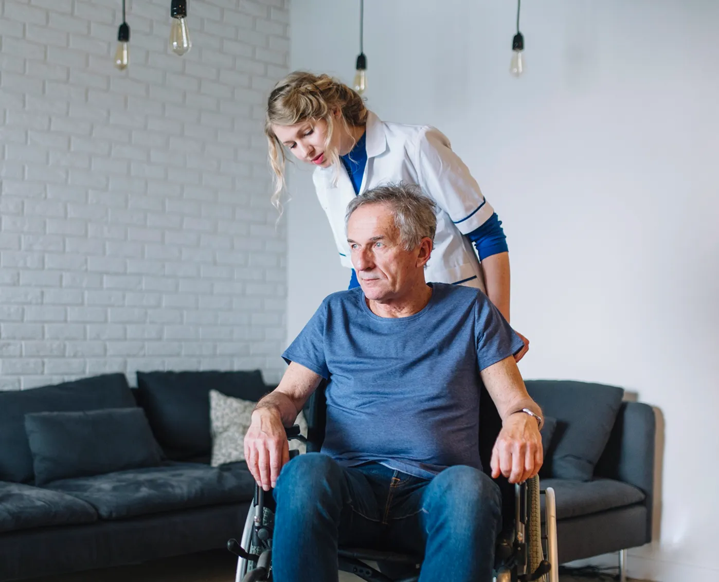 Caregiver stands behind an older man sitting in a wheelchair in a living room.