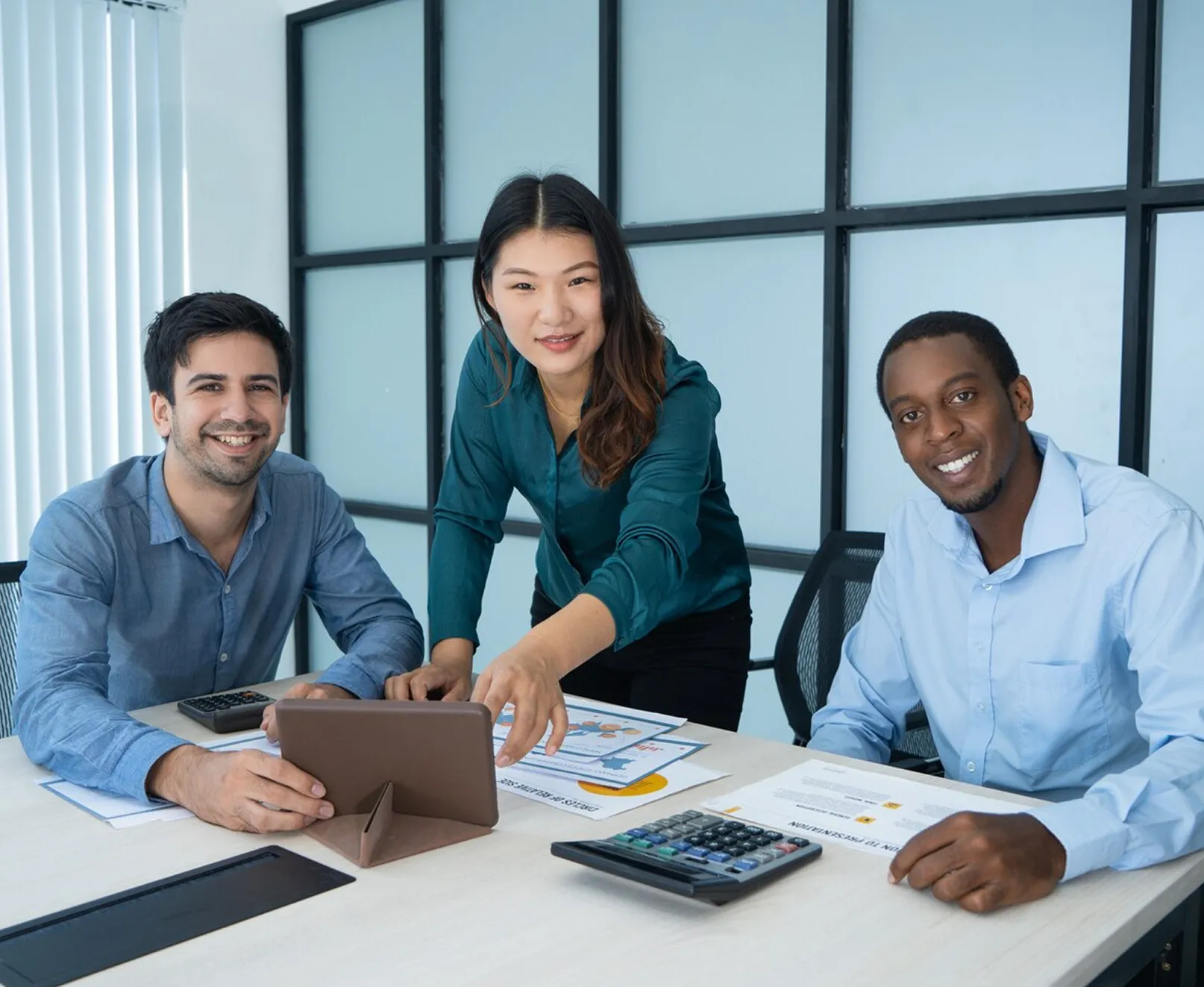 Three business people smiling at a table with a tablet, papers, calculators in a meeting room.