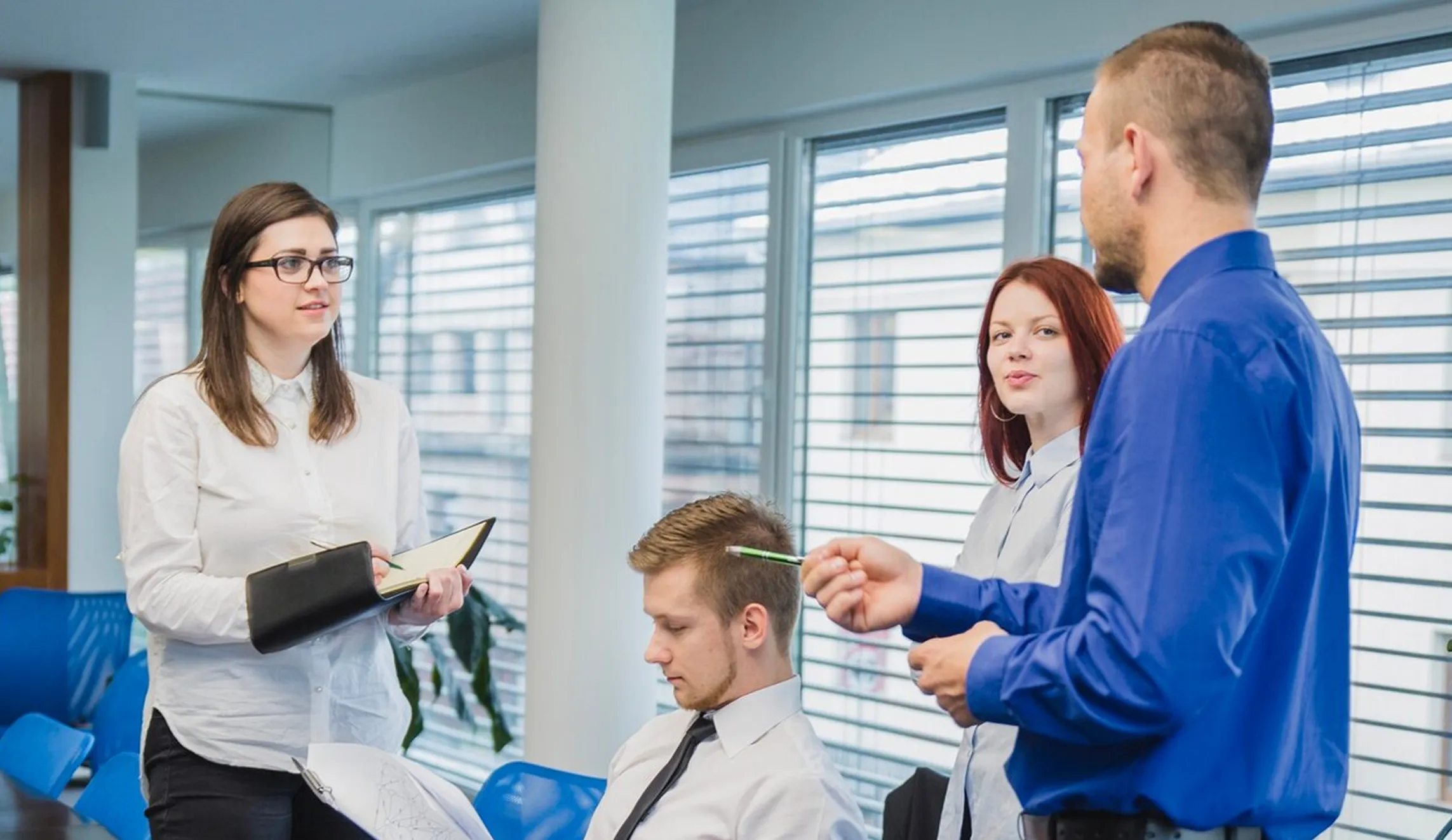 Four professionals in a meeting room having a discussion, one taking notes in a notebook.