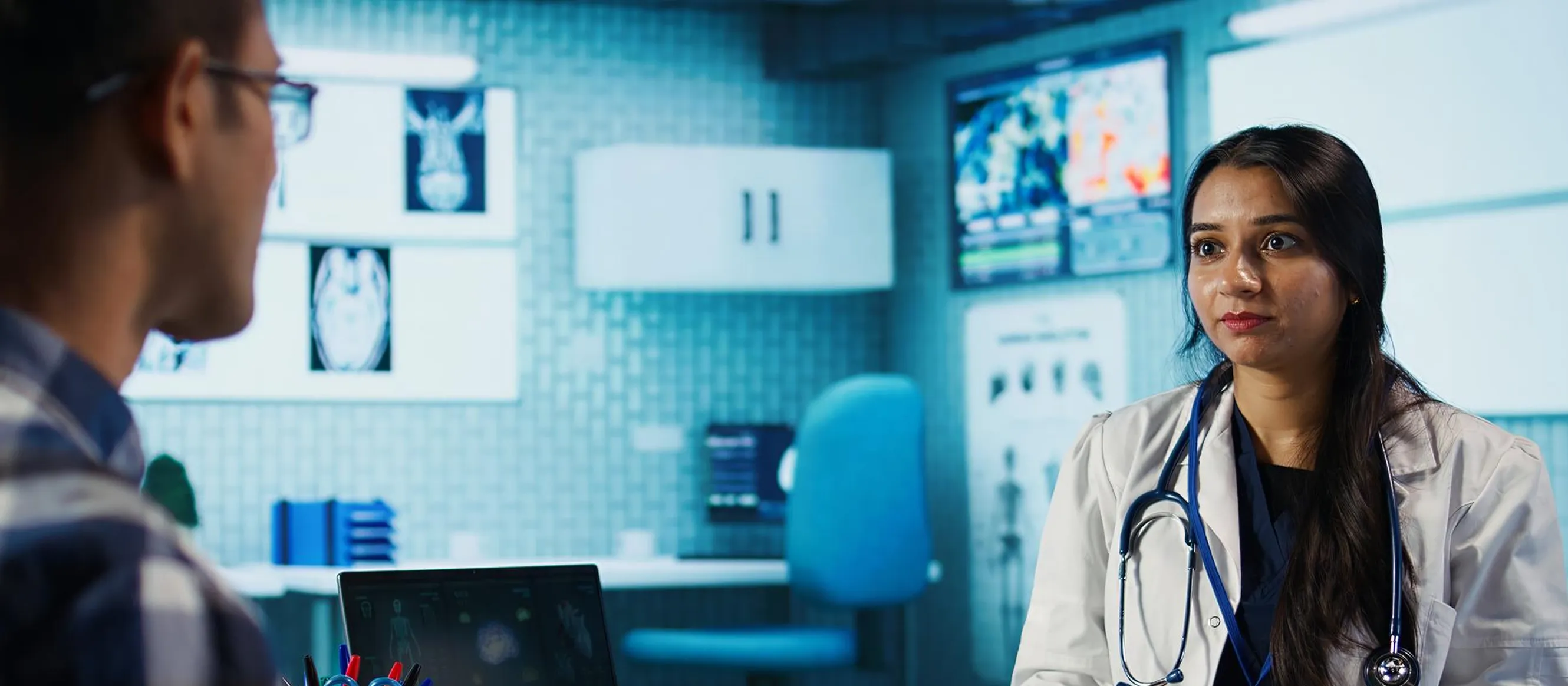 Female doctor with stethoscope talking to a patient in a medical office with scans on the screens.