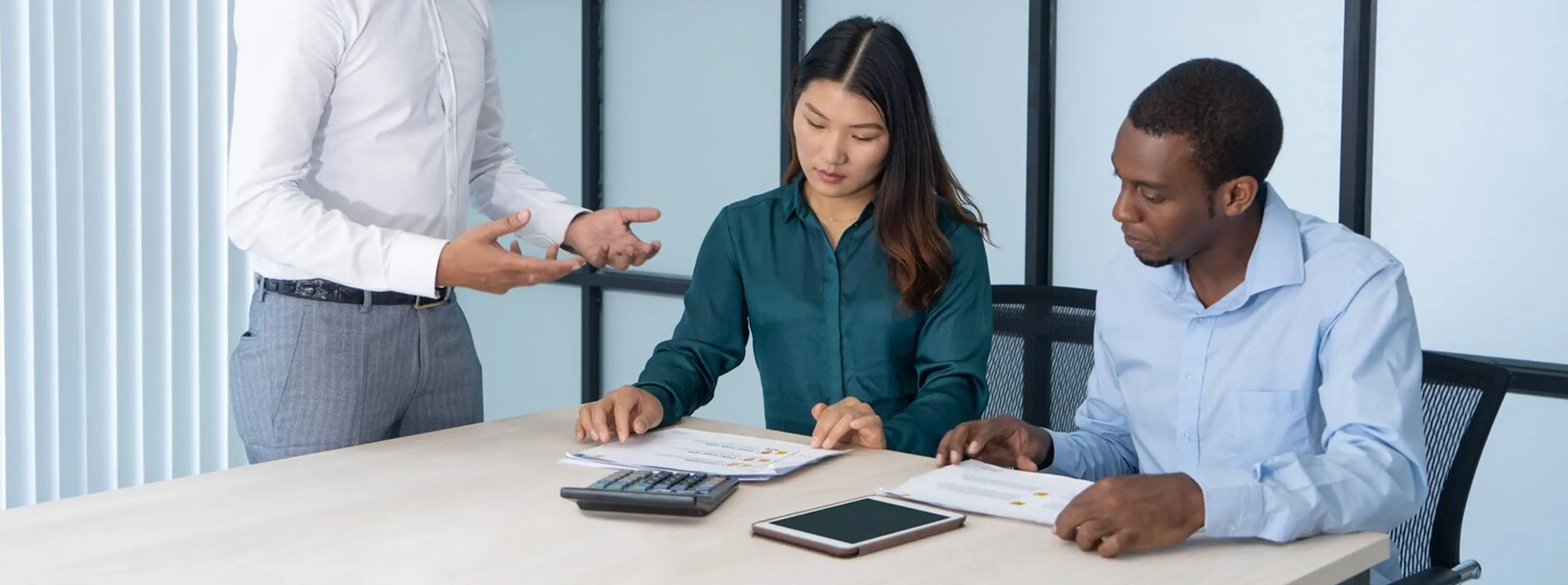 Two seated people reviewing documents at a table while a standing person gestures nearby.