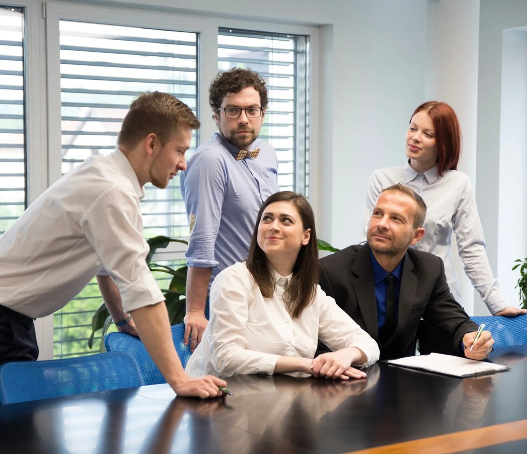 Five businesspeople gathered around a table discussing, with papers and pens in an office.