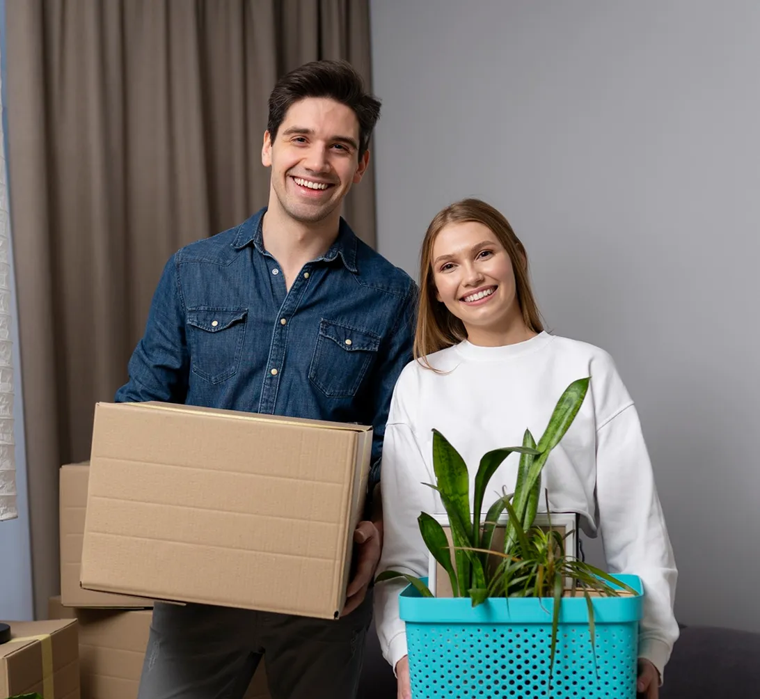 Smiling couple holding cardboard box and basket with plants, standing in a room with unpacked boxes.