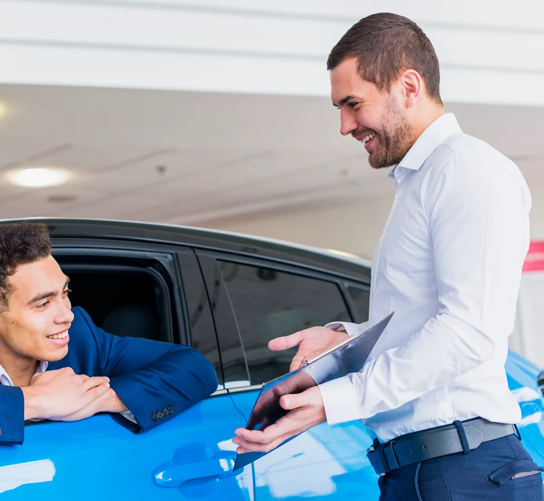 Car salesman in white shirt showing a clipboard to a smiling customer in a blue car.