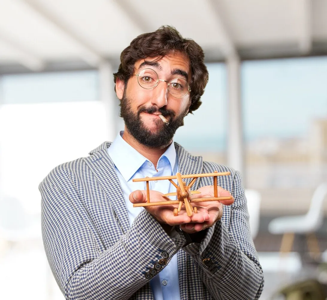 Bearded man with glasses pretending to smoke holding a wooden toy airplane in both hands.