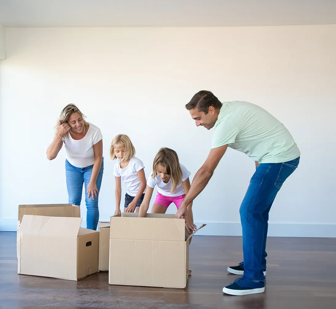 Family of four unpacking cardboard boxes in an empty room with wooden floor and white walls.